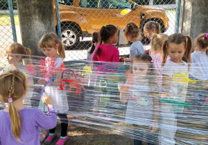 Średniaki malują farbami na foli streczowej rozwieszonej pomiędzy drzewami w ogrodzie przedszkolnym